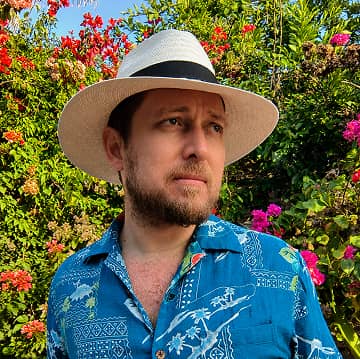 Portrait of Sebastián Iturralde wearing a white sun hat and blue tropical shirt, standing in a colorful flower garden outdoors.